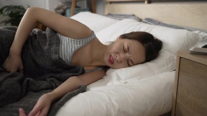 侧视亚洲妇女躺在白色床单上胃痛。生病的女士在家里舒适的地方睡觉时抱着痛苦的肚子。患病女性在卧室里腹部