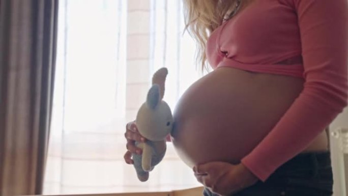 孕妇玩可爱的针织兔子玩具，抚摸她怀孕的肚子。快乐的妈妈准备好见到她心爱的男婴，等待孩子到来的女人。快