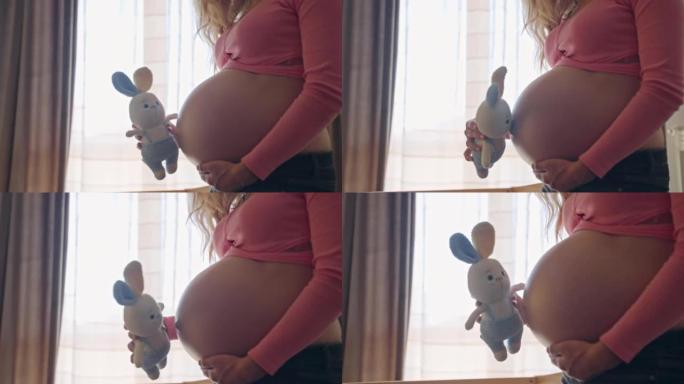 孕妇玩可爱的针织兔子玩具，抚摸她怀孕的肚子。快乐的妈妈准备好见到她心爱的男婴，等待孩子到来的女人。快