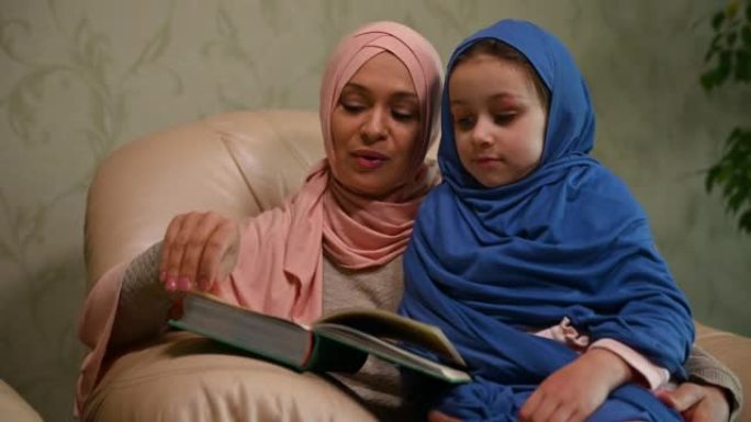 穿着粉红色头巾的穆斯林爱心妈妈为她的小女儿读了一本书，拥抱着她坐在现代客厅的沙发上