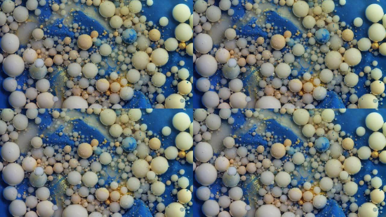 彩色金色丙烯酸涂料气泡散布在蓝色表面混合在抽象设计中。闪闪发光的灰尘，墨球滴和混合。运动中的多色液体