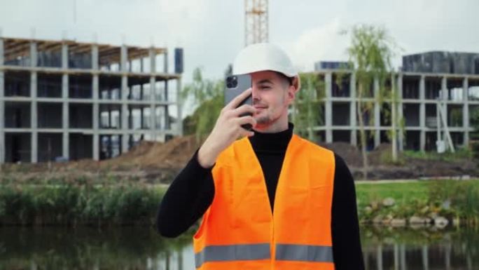 戴着白色安全帽的年轻迷人工程师用手机拍摄在建建筑的照片。男性通过智能手机向客户发送消息。工程项目。