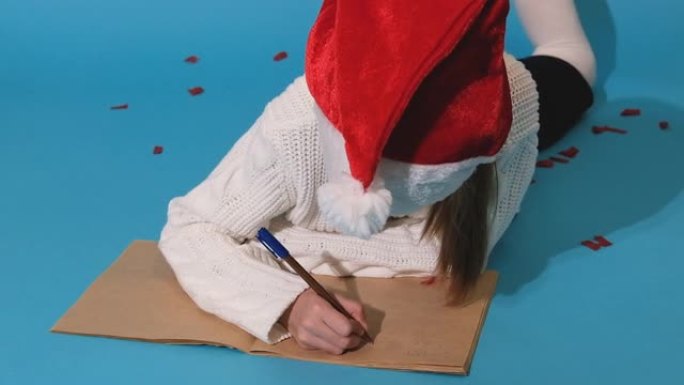 圣诞奇迹愿望清单。戴着圣诞老人帽子和白色毛衣的微笑女孩写信给圣诞老人的礼物。孩子躺在蓝色背景上。