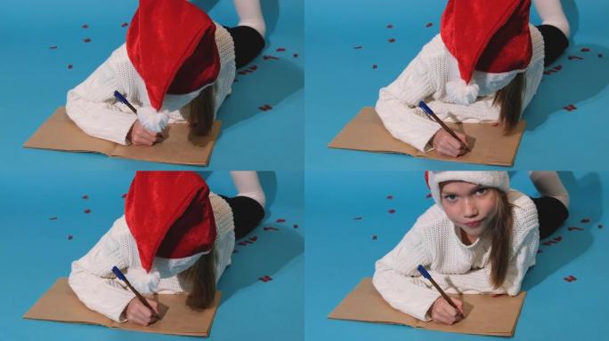 圣诞奇迹愿望清单。戴着圣诞老人帽子和白色毛衣的微笑女孩写信给圣诞老人的礼物。孩子躺在蓝色背景上。