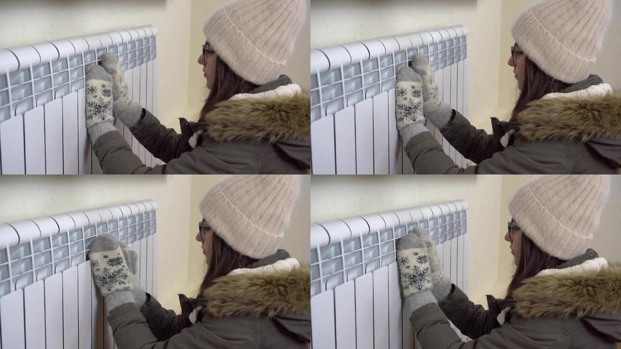 一个穿着冬季夹克，帽子和连指手套的年轻女孩用冲洗器温暖了双手。由于禁止房屋内的高暖气，房屋内很冷。4