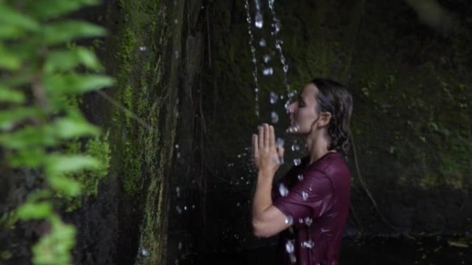 女人穿着湿衣服在森林岩石的天然小瀑布下冥想;获得能量、提神、放松和灵感。一个美女在瀑布边的慢镜头。