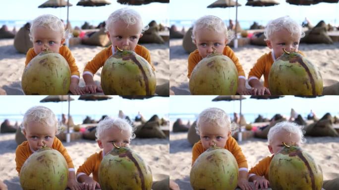 两个双胞胎幼儿喜欢在阳光明媚的海滩上喝新鲜的椰子，背景是草伞和海洋。与热带地区的孩子一起度假，以恢复