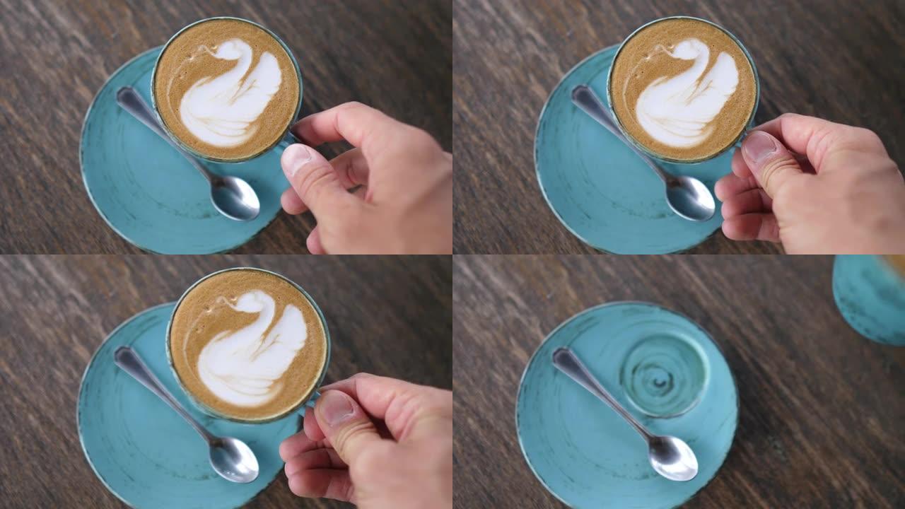 一个人的手的俯视图是从站在木桌上的碟子上举起的，是一杯植物性拿铁咖啡，泡沫上有拿铁艺术天鹅。纯素食饮