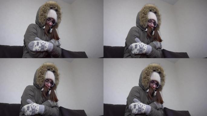 一个穿着冬衣的年轻女子坐在公寓里冻得要命。因为关了暖气，屋里很冷。戴眼镜、帽子、夹克和手套的女孩。特