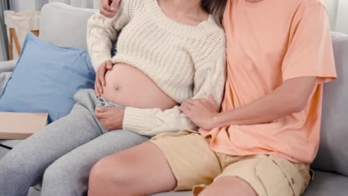 4K，近距离身体区域看不到这对夫妇的脸在沙发上拥抱，因为孕妇用手摩擦肚子，表现出爱意并等待即将到来的