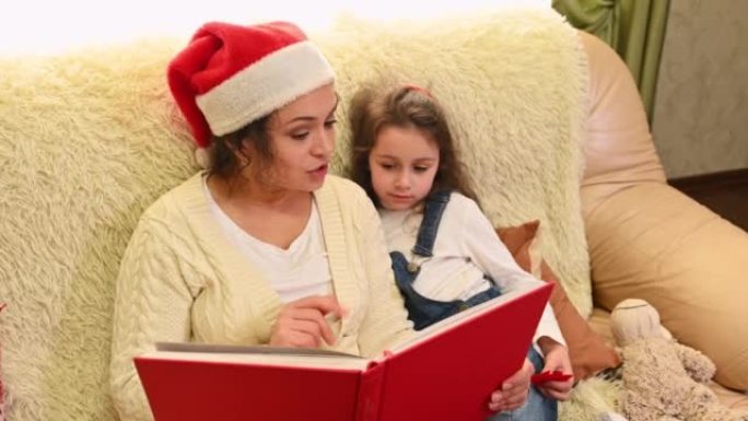 多民族的漂亮女人，戴着圣诞老人帽子的慈爱妈妈，坐在沙发上，给可爱的女儿读童话故事。