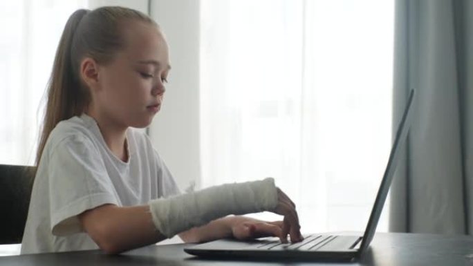 坐在灯光室桌子上的笔记本电脑上，用白色石膏绷带包裹着断手的可爱小女孩的侧视图。