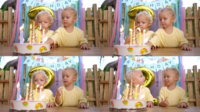 双胞胎婴儿正等着被吹灭生日蛋糕上的蜡烛。他们用手轻轻触摸生日蛋糕，然后将其从手指上舔下来。