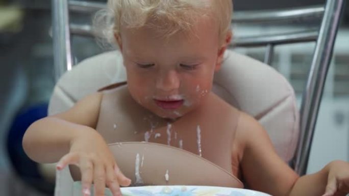 快乐的蹒跚学步的婴儿学会自己吃饭，在桌子上撒饭，手指弄脏，可爱的小孩脏脸，小孩吃牛奶粥。