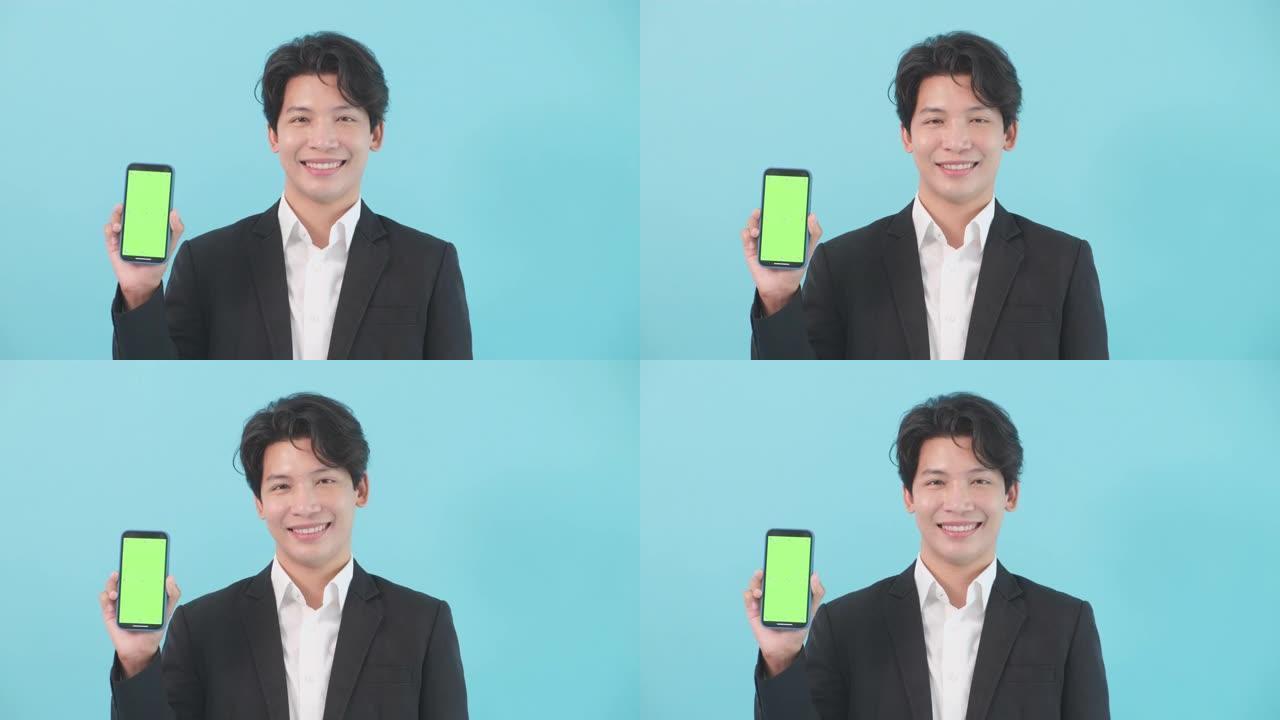 快乐的亚洲男人穿着西装，商人，拿着手机屏幕空白微笑看着相机，亚洲男人站在孤立的蓝色背景上拿着智能手机