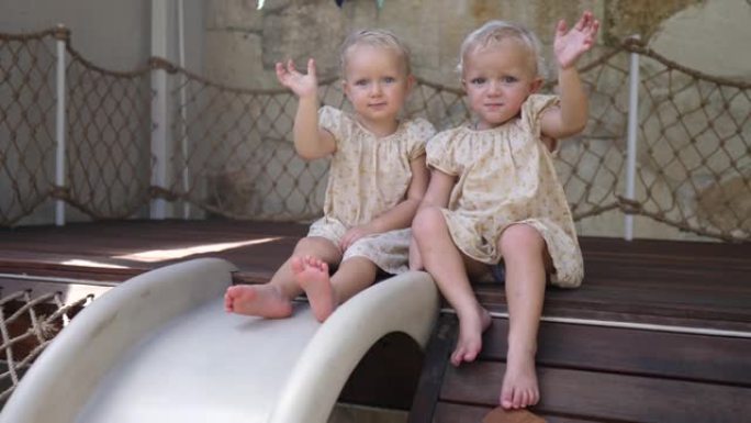 穿着黄色相配连衣裙的双胞胎女孩坐在儿童滑梯的边缘，准备滑下滑下并向相机挥手，以捕捉无忧无虑的童年。在
