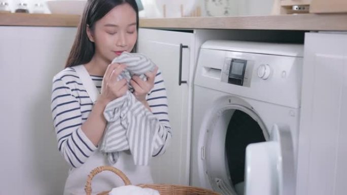 年轻快乐的亚洲女人在家里闻起来洗衣服。美丽的微笑女孩把衣服放在前面装洗衣机。家务和家务概念