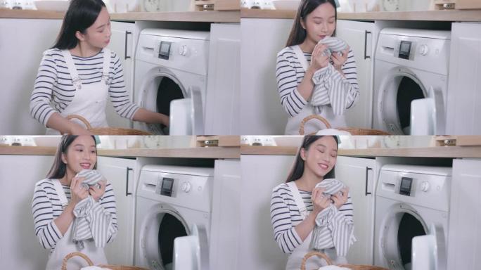 年轻快乐的亚洲女人在家里闻起来洗衣服。美丽的微笑女孩把衣服放在前面装洗衣机。家务和家务概念