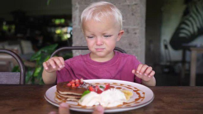 一个可爱的小疲倦的高加索孩子不想在为她服务的盘子上吃带有水果和奶油的煎饼。这孩子淘气，擦着眼睛。