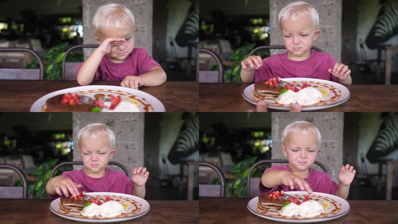 一个可爱的小疲倦的高加索孩子不想在为她服务的盘子上吃带有水果和奶油的煎饼。这孩子淘气，擦着眼睛。