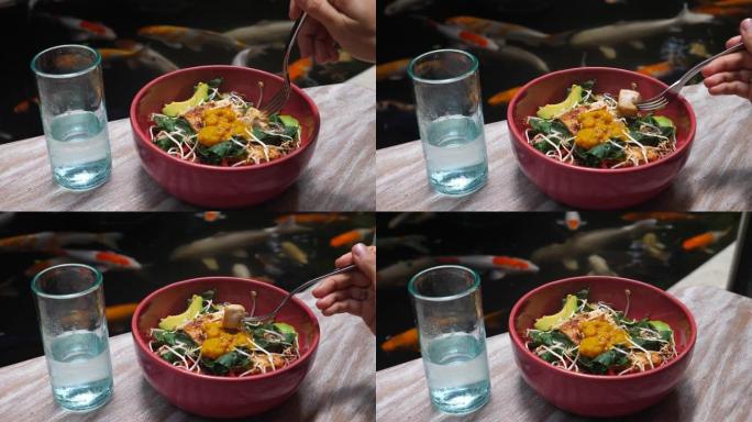 用叉子的手将健康的素食沙拉中的一块豆腐放在碗里。这顿饭在舒适的餐厅供应。桌子可以直接看到锦鲤池塘。