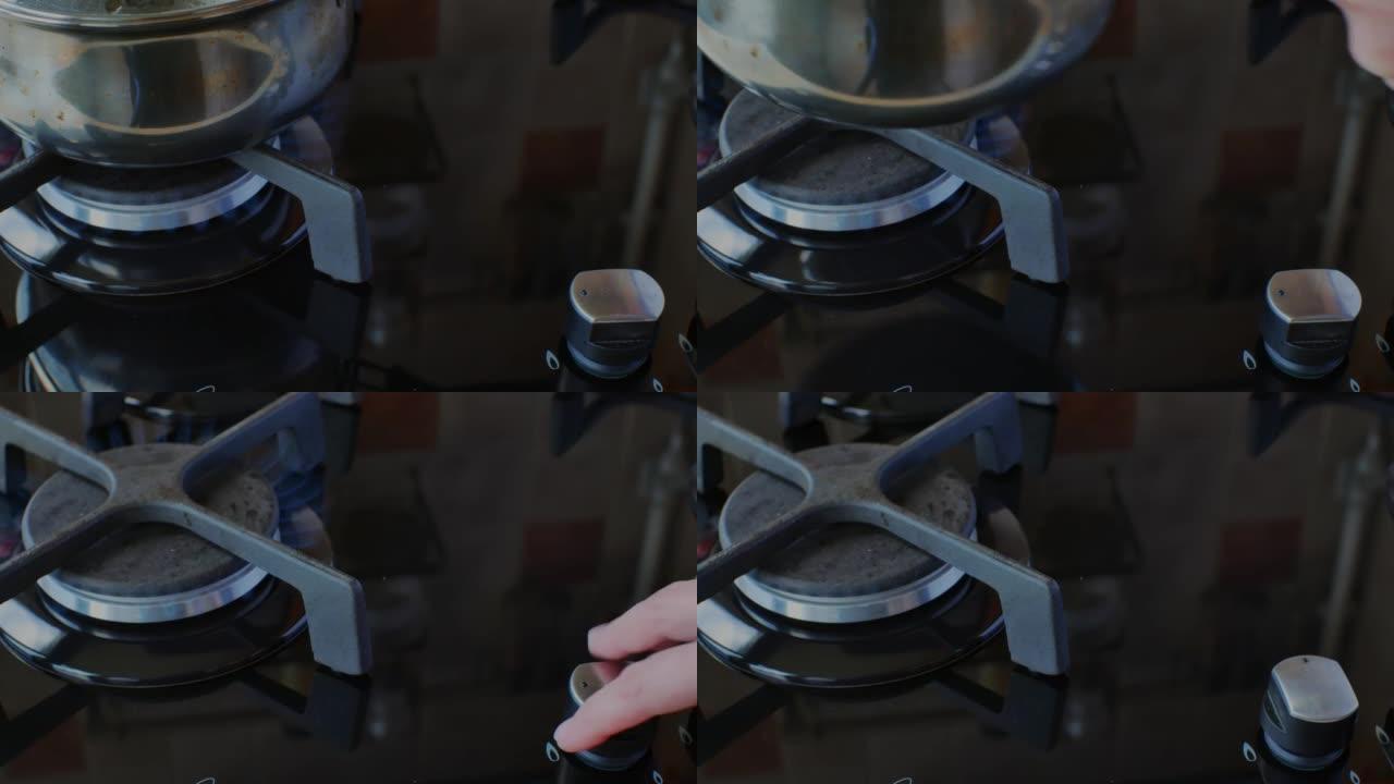 一只手的特写镜头，通过转动燃气灶上的旋钮并从炉子上取下不锈钢锅来关闭燃气。在现代厨房中使用天然燃料进