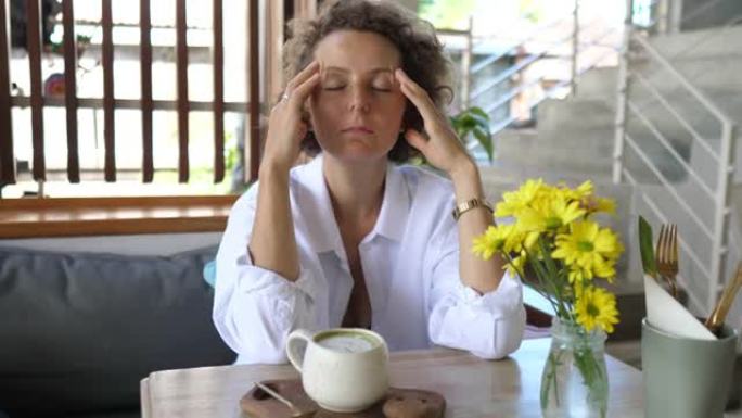 一位年轻的沉思白人妇女独自坐在咖啡馆里，桌上放着一杯抹茶。她摩擦太阳穴以缓解疼痛的头痛。精神卫生概念