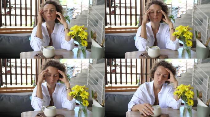一位年轻的沉思白人妇女独自坐在咖啡馆里，桌上放着一杯抹茶。她摩擦太阳穴以缓解疼痛的头痛。精神卫生概念