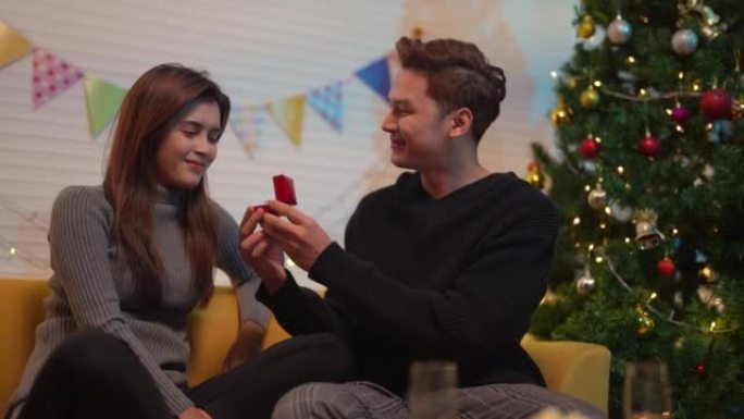 亚洲夫妇坐在沙发上，在客厅为婚礼送戒指。女人和男人在圣诞节或新年的长周末休息。圣诞主题的房间装饰庆祝