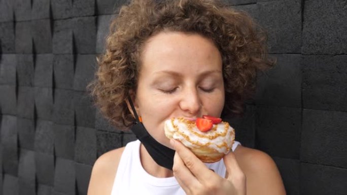年轻女子摘下口罩，吃一口美味的素食甜甜圈，上面放上切片草莓。口罩干扰吃美味的饭菜，在日常生活中使用防