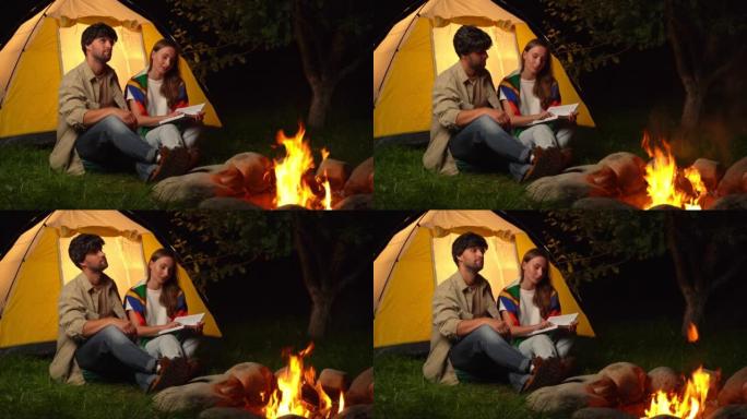 已婚夫妇晚上在篝火旁看书，以旅游帐篷为背景
