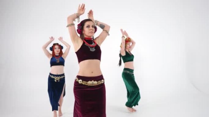 三个苗条的女人在慢动作中旋转，在白色背景下摇动臀部和胸部。自信的有才华的高加索肚皮舞者表演中东舞蹈。