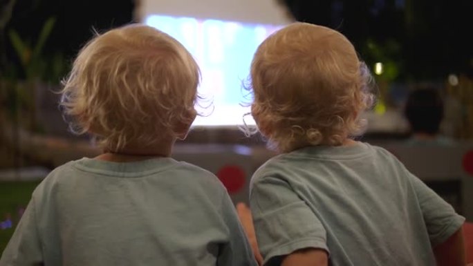 两个小金发女郎坐在沙发上，在电视大屏幕上观看儿童动画片的后视图。由于故事和绘画人物，儿童发展思维，逻