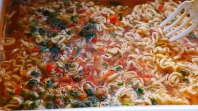 用叉子在一次性容器中搅拌特写煮熟的方便面。开胃的中国面条汤已经准备好了。选择性聚焦。