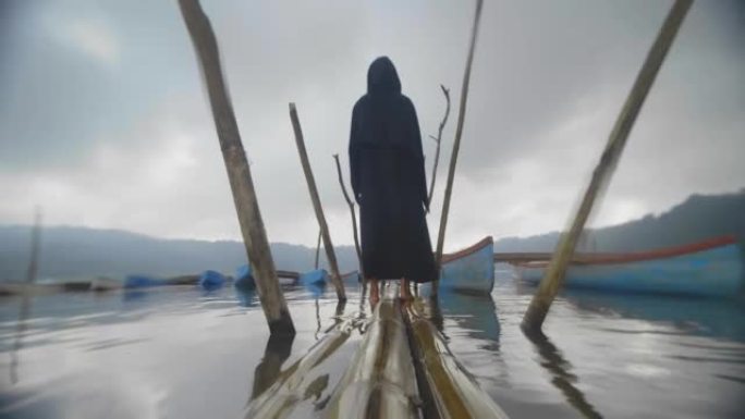 黑色兜帽男子站在热带湖上漂浮的旧码头上的背景图。实时