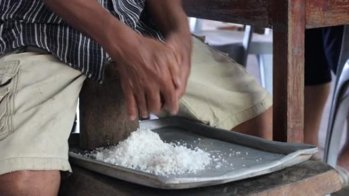 一位泰国老人坐在木椅上的特写镜头，用双手刮去白色的椰子肉。