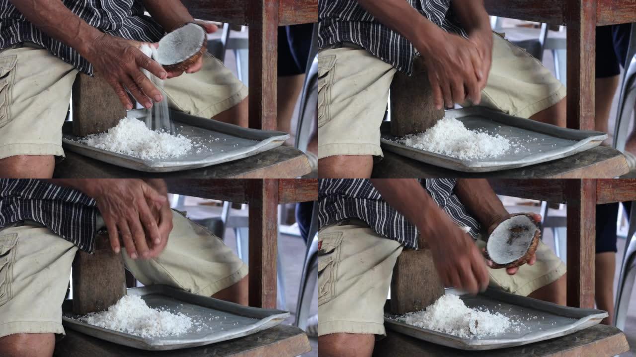 一位泰国老人坐在木椅上的特写镜头，用双手刮去白色的椰子肉。