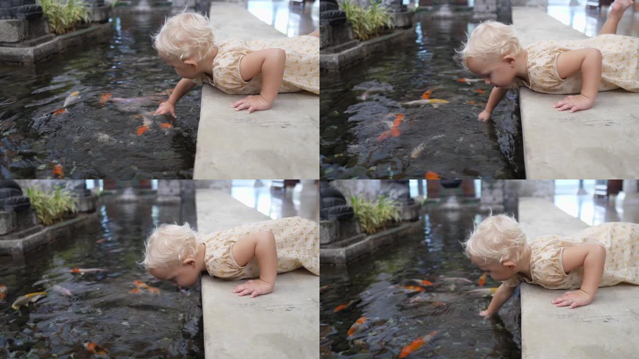 高加索2岁女孩躺在与水接壤的石阶上，在池塘里玩锦鲤鱼。孩子在水中移动他的手，发酵鱼的兴趣。