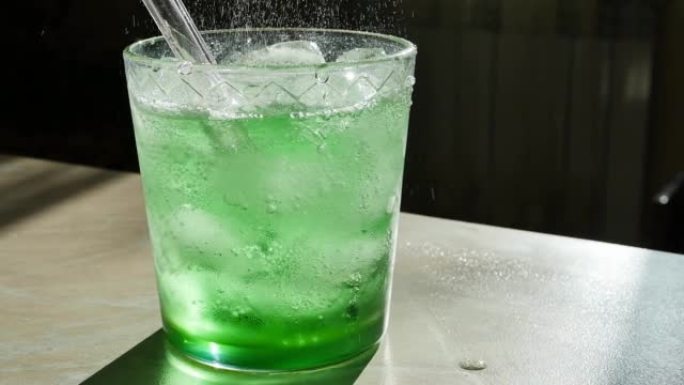 将绿色碳酸饮料倒入装有冰块和可重复使用的玻璃吸管的玻璃杯中，以慢动作多莉射击。龙蒿或苦艾酒的流动和飞