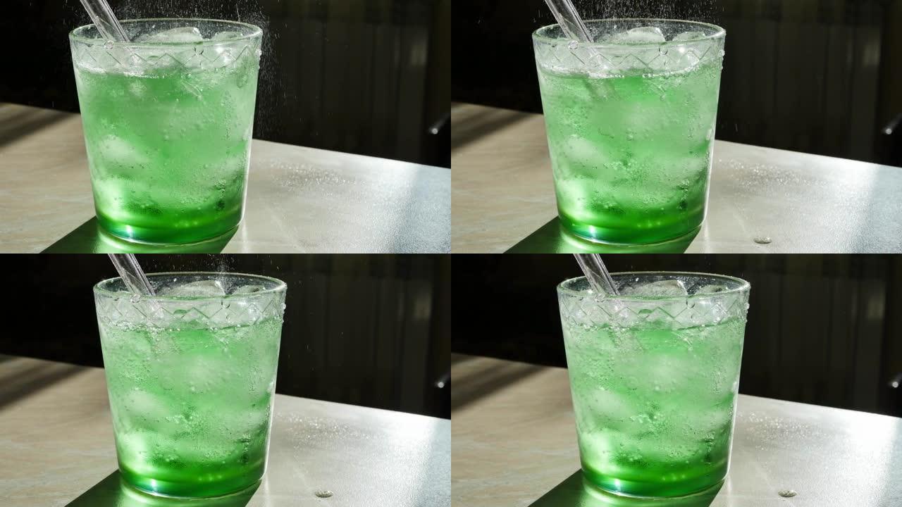 将绿色碳酸饮料倒入装有冰块和可重复使用的玻璃吸管的玻璃杯中，以慢动作多莉射击。龙蒿或苦艾酒的流动和飞