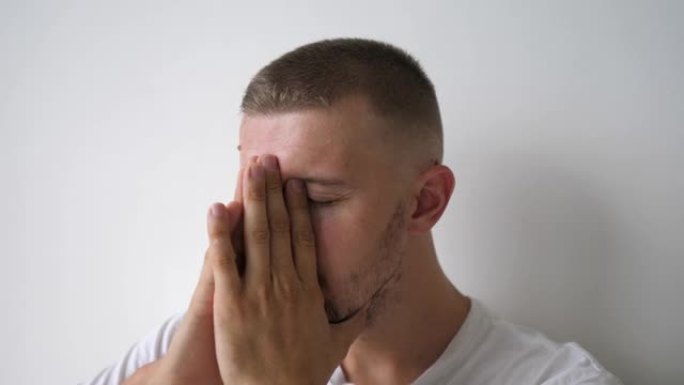 一个年轻的高加索人坐在白色的墙上，用手遮住脸。他正在经历失去亲人的痛苦，并试图应对自己的抑郁状态。