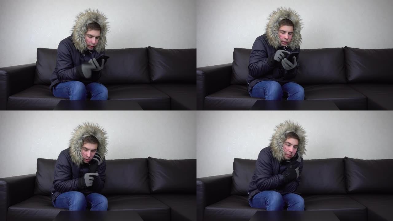 一个年轻人穿着冬季夹克坐在家里，试图保暖。一名男子坐在没有暖气的情况下，打电话寻求帮助。4k