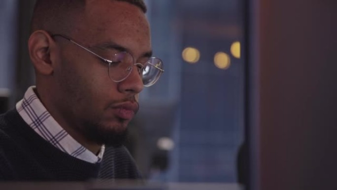 戴着眼镜的年轻非洲裔美国人坐在现代的联合办公办公室里，在电脑前工作