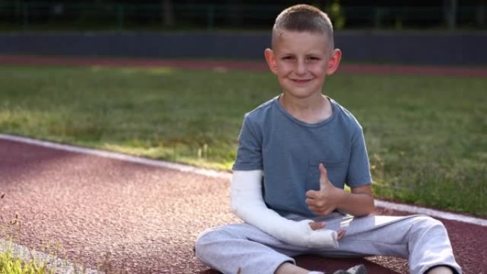 夏天，微笑的断手小男孩坐在户外运动场上。9岁的孩子，四肢骨折，显示另一只健康的手，拇指向上。手臂上的