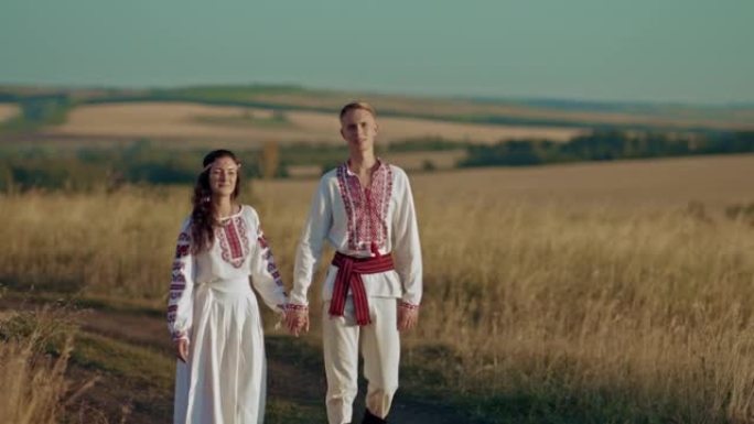 美丽的男人和一个穿着乌克兰民族服装的女人正沿着一条乡村道路行走，他们很高兴。男人和女人手牵手，人民的
