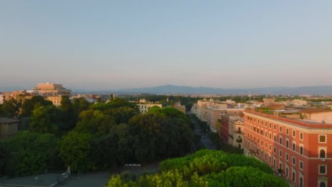 公园中生长的树木的鸟瞰图在低太阳照亮的公寓楼之间。向前飞行在市区上空。意大利罗马