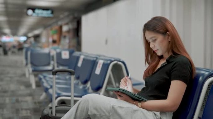 年轻的亚洲妇女在机场等待航班时在数字平板电脑上工作。