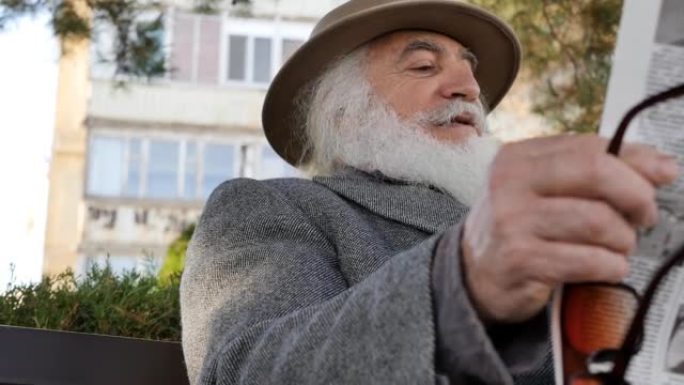一个留着胡须，戴着帽子的老人坐在公园的长椅上看报纸。一位大胡子的老人在公园放松时阅读报纸上的最新新闻