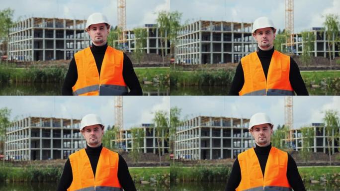 建筑工地的专业男性工业工人。帅气的工业工人看着相机穿着高高的背心，戴着安全帽头盔，在盖房子的背景下拿