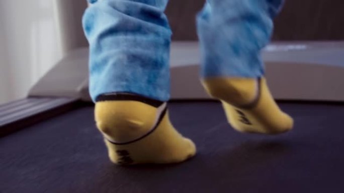 一个穿着牛仔裤和黄色袜子的孩子在跑步机上行走。在家运动